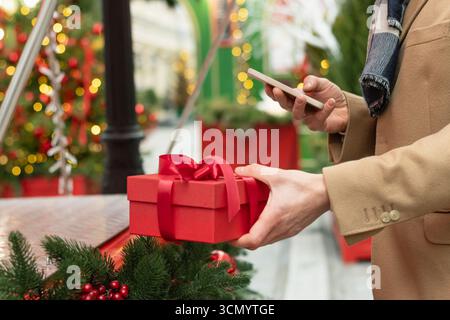 Mans hand holding red gift box as present adorned and using smartphone. Christmas fairy. Xmas shopping. Side view. Close up. Stock Photo