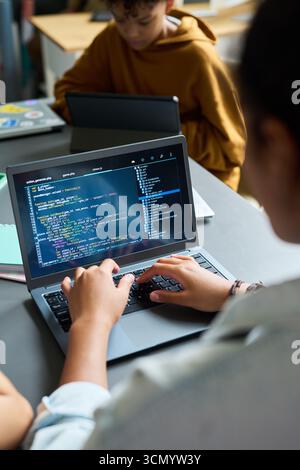 Teenager typing code on laptop, sitting at desk in classroom, another teenager with curly hair using laptop in background, focusing on programming activity Stock Photo
