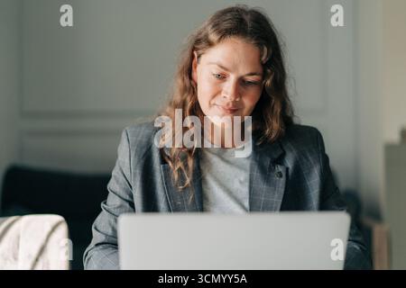 A middle-aged woman sitting at home in a business suit works on a laptop. Stock Photo