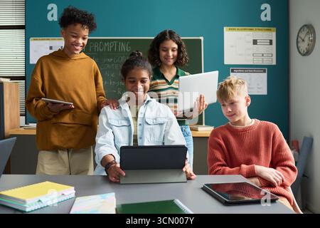 Group of multiethnic teenagers collaborating on digital tablets and laptop in classroom, smiling and interacting with technology, diverse students engaging in educational activity together Stock Photo