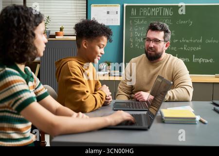 Caucasian man teaching multiethnic teenagers using laptops in classroom, students listening and engaging in discussion, chalkboard with AI prompting instructions visible in background Stock Photo