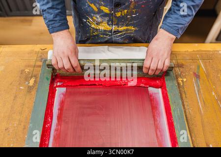 A first-person view of a craftsman printing on fabric with red ink in a screen printing workshop. A symbol of creativity and manual work. Stock Photo