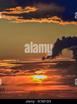 Dramatic sunset sky showing bold fiery tones fading into cold grays, creating magical atmosphere with hidden sunlight behind clouds. Stock Photo