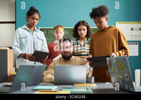 Caucasian man teaching diverse group of teenagers using digital tablets and laptops in classroom, students standing around teacher engaging with technology and learning Stock Photo