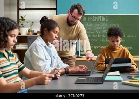 Caucasian man instructing multiethnic teenagers using laptops and tablets in classroom, students focusing on digital devices while teacher providing guidance near chalkboard Stock Photo