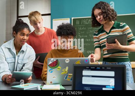 Group of multiethnic teenagers collaborating on digital project in classroom, using laptop and tablets, standing and sitting around desk, focused on technology and teamwork Stock Photo