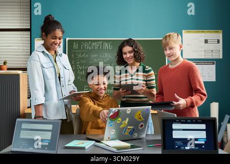 Group of multiethnic teenagers collaborating on coding project, smiling and interacting with laptop and tablets in classroom, digital devices and AI prompt instructions visible on chalkboard Stock Photo