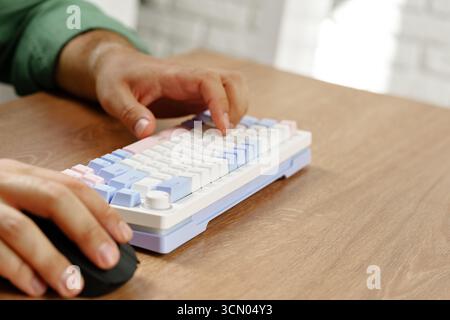 Hands typing on a pastel keyboard while using a computer mouse on a wooden desk Stock Photo