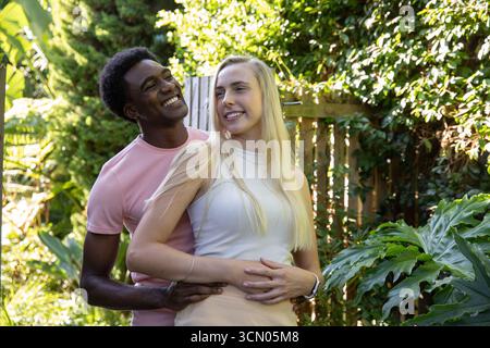 Diverse couple embracing in leafy backyard garden with smartwatch, wooden fence and tropical plants. Romance, outdoor, nature, tranquility, vitality, Stock Photo