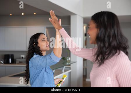 Diverse female friends giving high-five while using spirit level by sink and air fryer in kitchen. Collaboration, achievement, teamwork, celebration, Stock Photo
