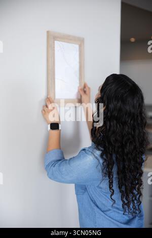 African American woman adjusting picture frame against wall at home wearing smartwatch, copy space. Minimalist, modern, interior, decor, lifestyle, de Stock Photo