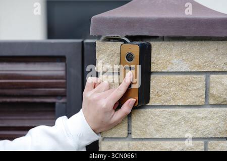 Woman using smart video intercom system, pressing button on electronic device installed on brick wall near entrance gate Stock Photo