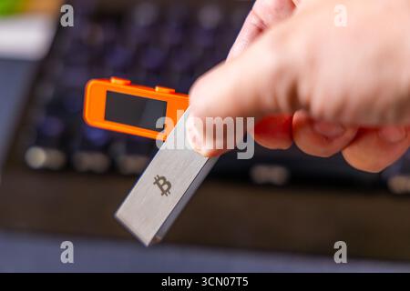 Adult man with light skin holds cryptocurrency hardware wallet in front of computer keyboard. Stock Photo
