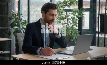 Pensive puzzled Caucasian business man businessman male entrepreneur corporate employee sales manager working in office typing on laptop searching Stock Photo