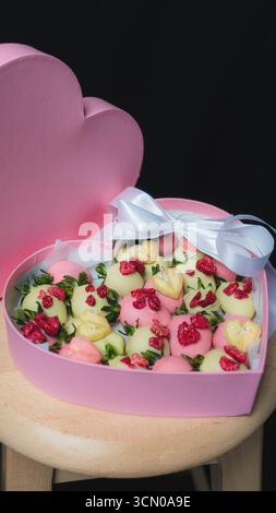 A heart shaped pink gift box filled with strawberries in white and pink chocolate, decorated with dried raspberries and green leaves. Elegant dessert Stock Photo