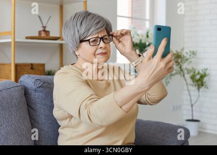 Senior woman sitting on sofa at home holding eyeglasses trying to read message on smartphone Stock Photo