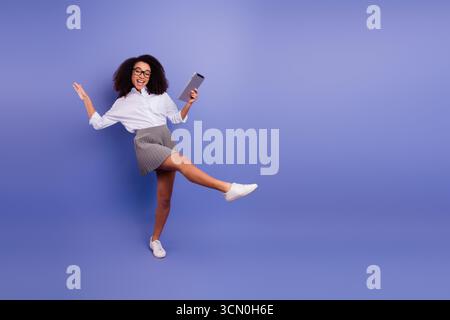 Cheerful young woman in formal attire posing joyfully against a colorful violet background while holding a digital tablet. Stock Photo