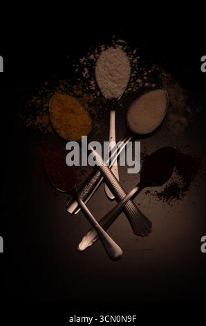 Five metal spoons with flour, sugar, and spices on a dark background, scattered powders creating a rustic, minimal gourmet food composition. Stock Photo