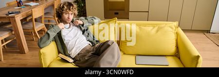 Young man in casual clothes sits on a bright yellow couch, looking relaxed and thoughtful. Stock Photo