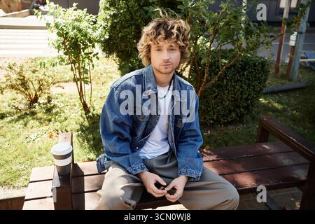 A stylish young man relaxes on a bench, sipping coffee under the warm sun in an urban park. Stock Photo