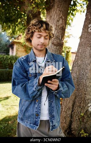A handsome man in casual attire enjoys writing in a notebook under a large tree in daylight. Stock Photo