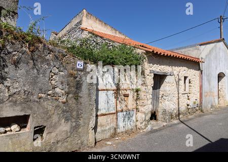An old, abandoned building in a rural Portuguese village features a rusted gate, a number plate "6", and a stone facade overgrown with plants. Stock Photo