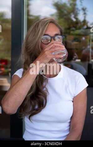 An older, active, athletic woman drinks a natural mineral water glass every day to stay fit and healthy. The pensioner wears glasses and laughs. Stock Photo