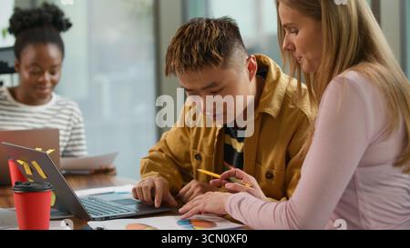 Informal casual business team three diverse multiracial colleagues working together at office Asian businessman helping Caucasian female analyze Stock Photo