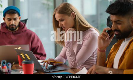 Three concentrated business professional multiracial colleagues at table informal working company busy job task Caucasian woman with laptop Indian men Stock Photo