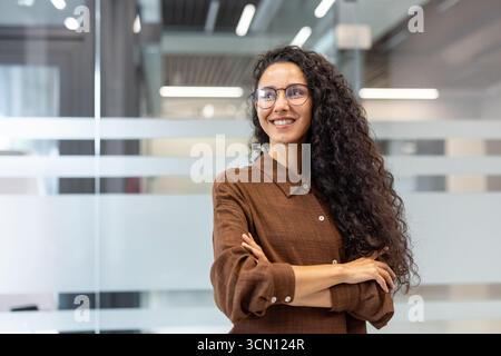 Confident young woman with curly hair and glasses standing in a contemporary office setting, crossing arms and looking away, representing success and professionalism in business Stock Photo