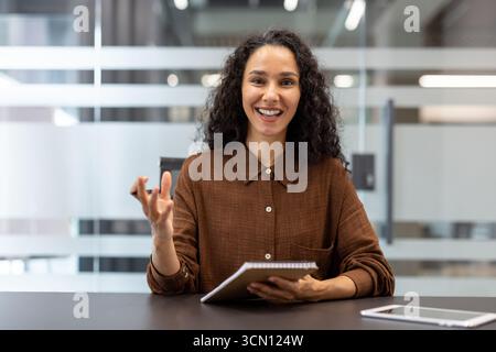 Young professional businesswoman smiling, looking at camera and gesturing, participating in a virtual meeting from a modern office workspace with a notebook Stock Photo