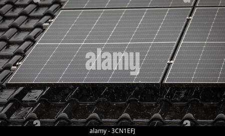 Black solar panel on glossy black roof tiles during heavy rain, raindrops splashing on panel, partial view of roof behind, 16:9 format Stock Photo