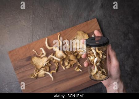 Hand holding glass jar with black lid filled with dried oyster mushrooms on dark slate, wooden board with mushrooms at bottom. Stock Photo