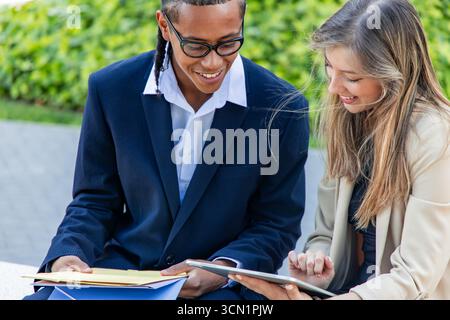 Multiethnic business partners smiling while reviewing documents and engaging with a tablet in the bustling financial district, showcasing teamwork and Stock Photo