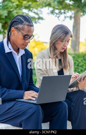 Two young business partners are working outdoors with a laptop and a digital tablet, collaborating on a project in the financial district, utilizing t Stock Photo