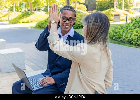 Two business partners celebrating a successful deal with a high five while working on a laptop in a vibrant financial district, showcasing teamwork an Stock Photo