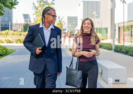 Multiethnic business partners are walking and talking in the financial district during their lunch break, enjoying the urban landscape and discussing Stock Photo