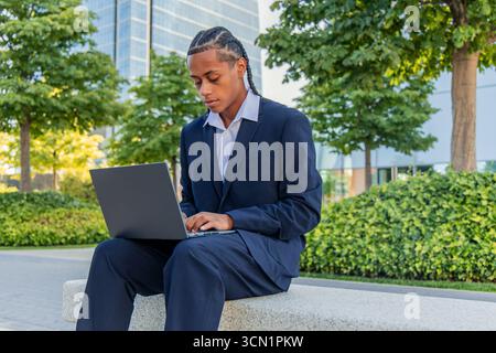 Young businessman sitting on a low wall in a tree-lined park within a bustling financial district, typing intently on his laptop while immersed in his Stock Photo