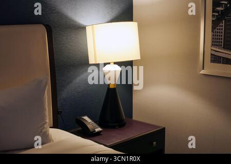Hotel room with bedside lamp. Night table and interior lighting. Modern accommodation furniture decor. Comfortable bedroom detail view. Stock Photo