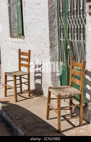 two vintage wooden chairs with wicker or rush seats either side of a doorway on the pavement outside of a mediterranean home casting shadow. Stock Photo