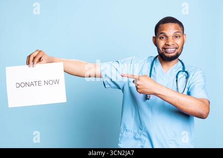 African american nurse advertising the message to donate blood, holding a paper and urging people to help in studio. Medical assistant in workwear standing against blue background. Stock Photo