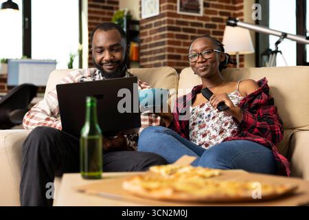 Smiling black man works remotely on laptop while girlfriend shares bowl of chips and controls TV with remote control. Happy couple enjoying relaxed weekend evening in cozy brick wall apartment. Stock Photo
