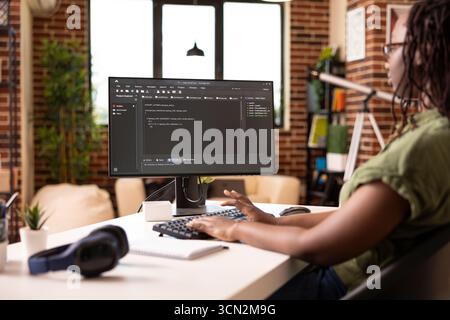 Black woman developer works from home, writing and troubleshooting code for tech startup. Female entrepreneur focused on app development using desktop pc in brick wall apartment. Stock Photo