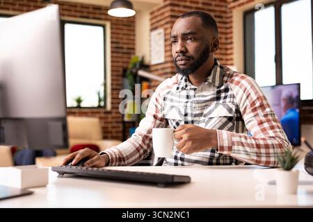 Self employed black man at home office, looking at desktop monitor with worried expression, reviewing company documents. Young male entrepreneur holding coffee mug and checking business financial data Stock Photo