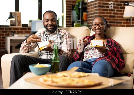 Happy male and female friends seated on couch holding takeout boxes, having lunch at home. African american couple having cozy weekend hangout with food and drinks in brick wall apartment. Stock Photo
