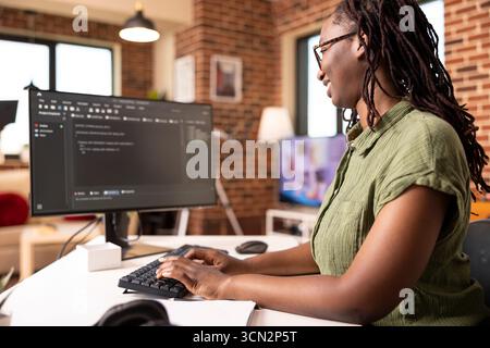 Black female software developer codes securely from home office, using computer to review programming scripts. African american IT freelancer working on tech solutions in modern apartment. Stock Photo