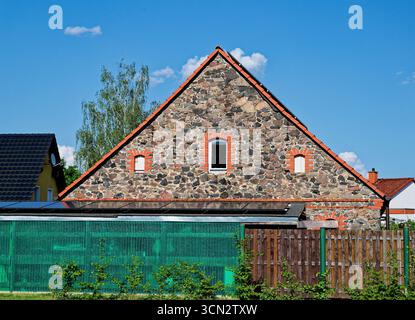 A two story house built from large stone blocks set in mortar shows a neat plastic window near the roof, framed by brick detail. A wooden fence and a Stock Photo