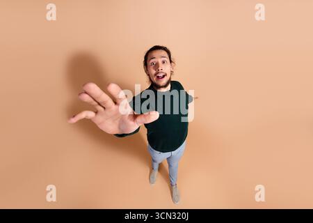 Expressive young man reaching forward with an engaging gesture against a beige backdrop showcasing a fashionable style Stock Photo