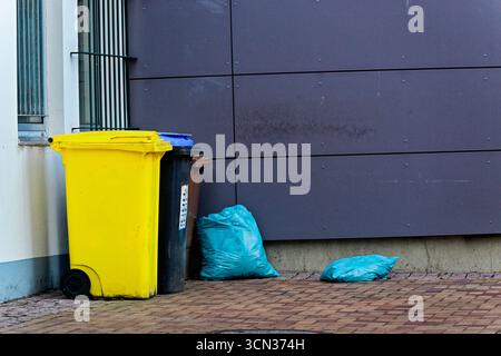 Three city bins stand together for plastic paper and organics. A blue bag and smaller pieces lie on the ground. A dark wall and a beige house with win Stock Photo