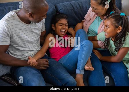 Diverse family tickling daughter while laughing in home living room on dark blue couch with remote Stock Photo
